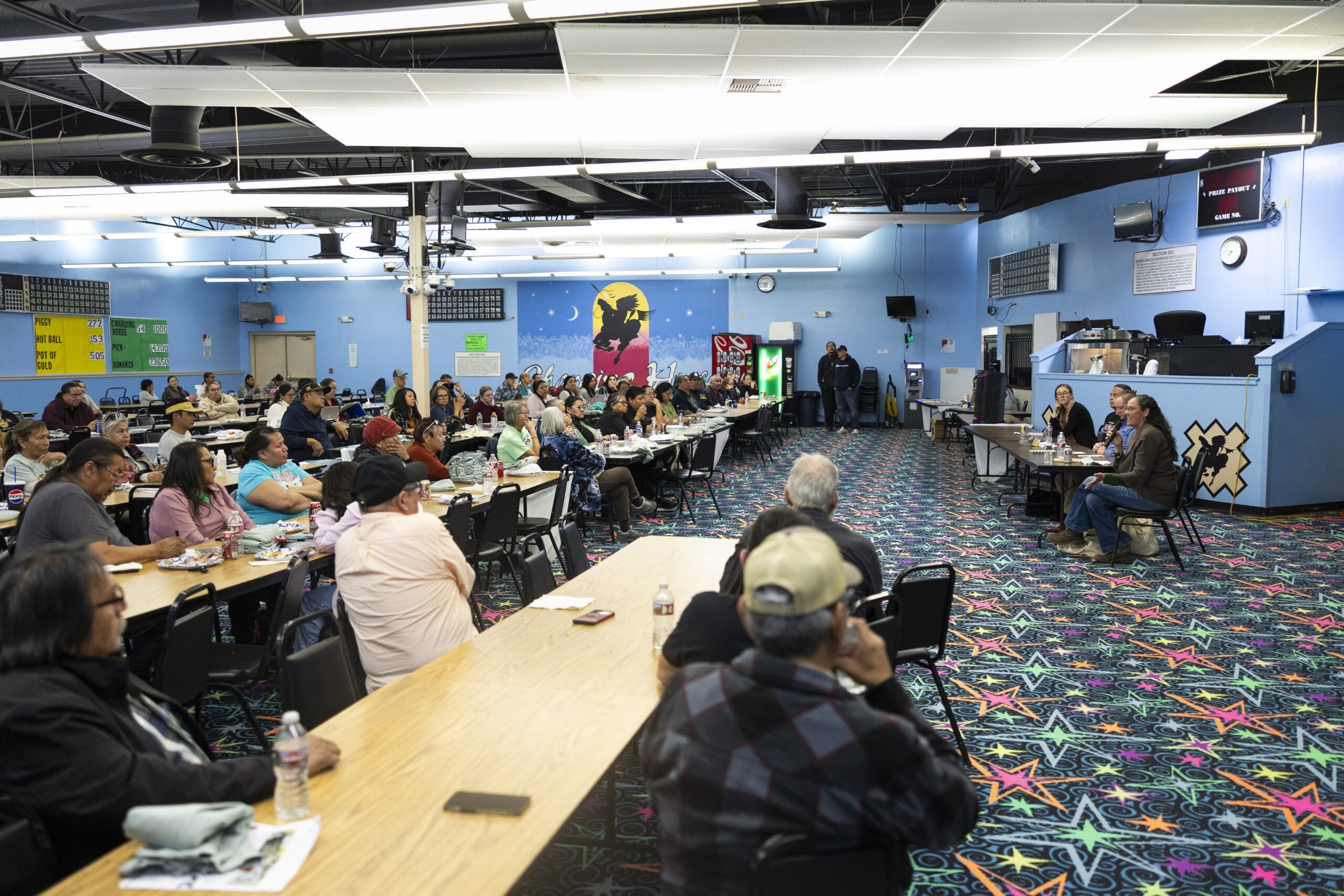 A wide interior shot of a community hall, with dozens of people seated at long tables, looking toward a panel of speakers at the front of the room.